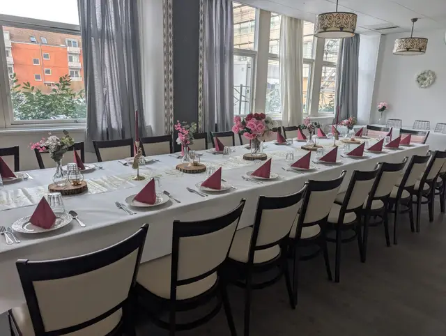 Salen dækket til fest: et langbord ved vinduerne med mørkerøde servietter og lyserøde blomster ved hver kuvert