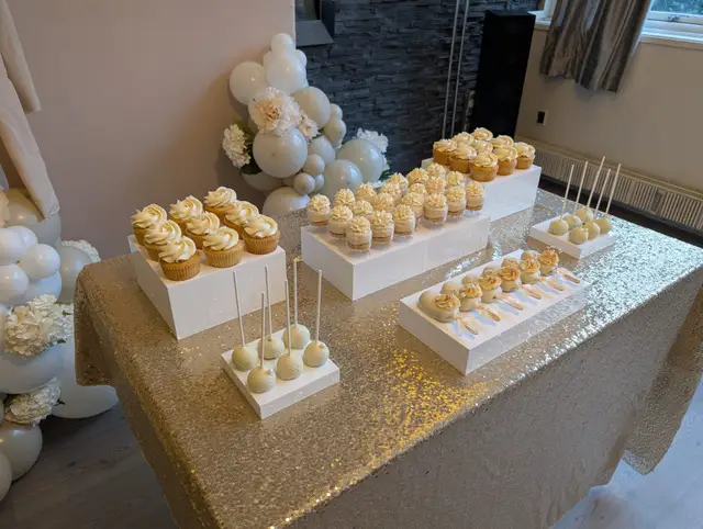 A dessert table of cupcakes and cake pops on white risers over a gold sequinned cloth, below a balloon arch